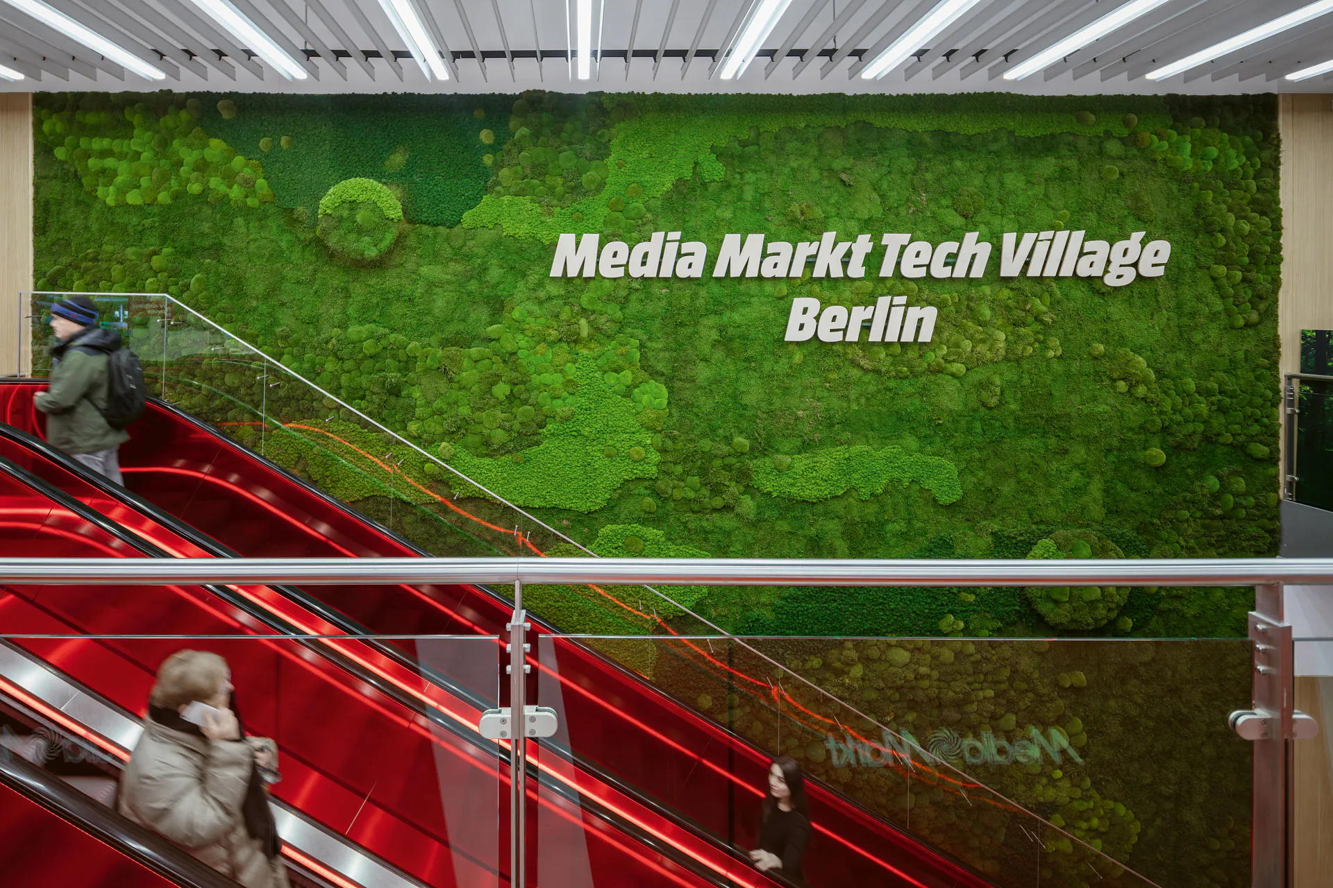 Großflächige grüne Moosswand mit weißer Beschriftung 'Media Markt Tech Village Berlin' in moderner Empfangshalle mit roten Treppen und LED-Deckenbeleuchtung.